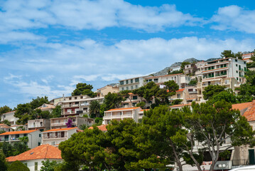Obraz premium Picturesque hillside town with houses featuring orange-tiled roofs and light-colored walls, surrounded by lush green trees under bright blue sky with scattered clouds. Croatia, Brella - June 4, 2006
