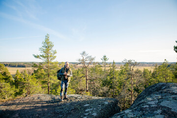 Male hiker taking photos with vintage camera on scenic cliff in Finland. Travel photography, adventure tourism and outdoor lifestyle concept.