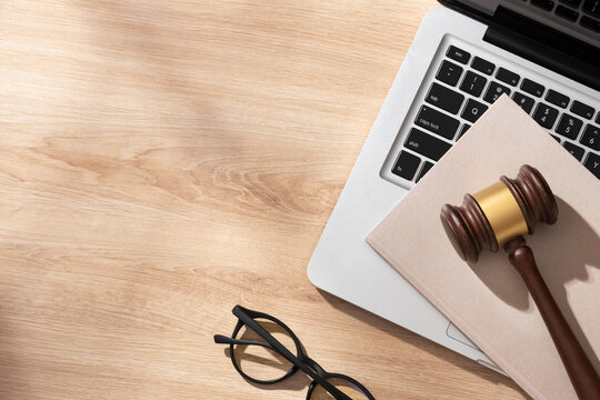 A wooden gavel with gold band sits on a light wooden desk beside a laptop, beige notebook and eyeglasses, evoking a modern legal workspace under soft lighting. Top view.