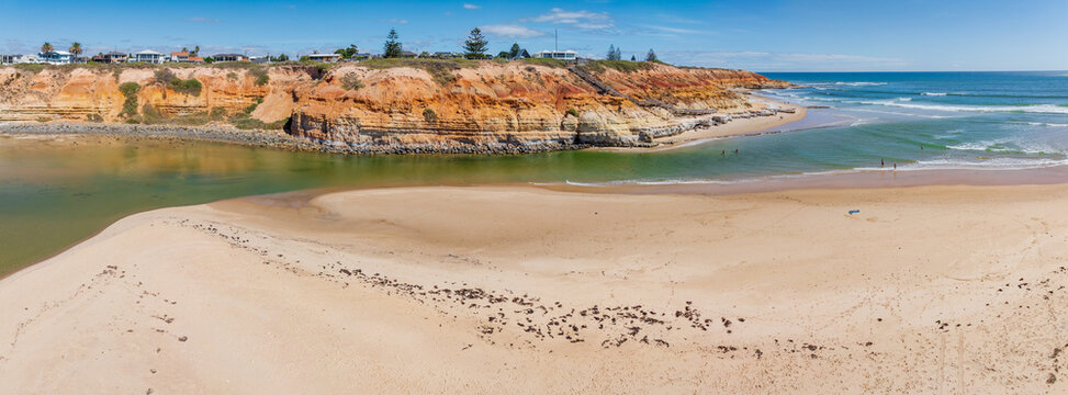 Low level aerial panorama of a coastal river flowing out to sea between a beach and a rugged cliff