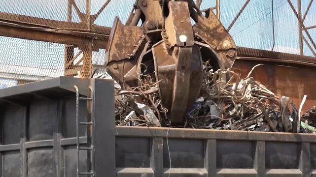 A large mechanical grapple crane efficiently transfers tangled piles of metal scrap and recycled iron into the open bed of a commercial transport truck at a busy industrial recycling facility
