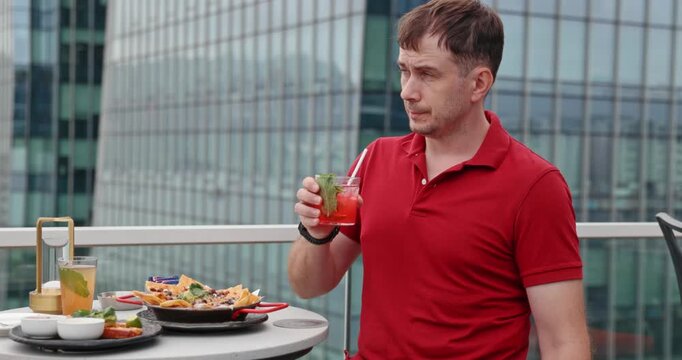 Man in red polo shirt drinking cocktail at rooftop bar with city skyscrapers background. Urban lifestyle, luxury dining, summer vacation, travel, relaxation, city break.