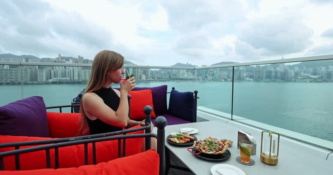 Woman enjoying a refreshing cocktail and delicious meal at a luxurious rooftop restaurant, gazing at the panoramic city skyline and harbor. Urban dining, luxury travel, leisure lifestyle, city break