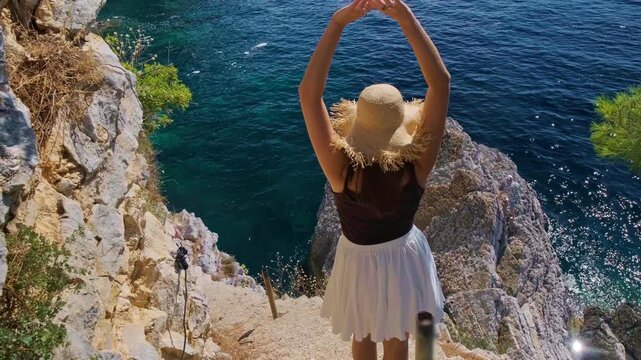Girl in straw hat standing back tossing white flowing skirt raising arms on cliff edge narrow path above blue Adriatic Sea Petrovac Montenegro