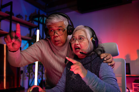 Senior couple, excited senior eldery Asian woman and man with headphones streamer playing video game using desktop computer at gaming room at home. colorful neon background