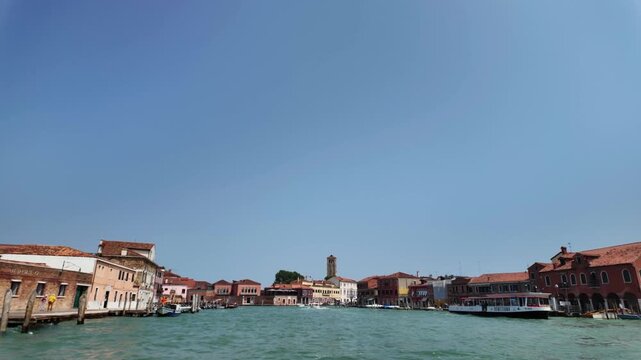 View of Venice from a boat slowly approaching across the lagoon. On either side, old houses built close to the water, boats, and pedestrian walkways.