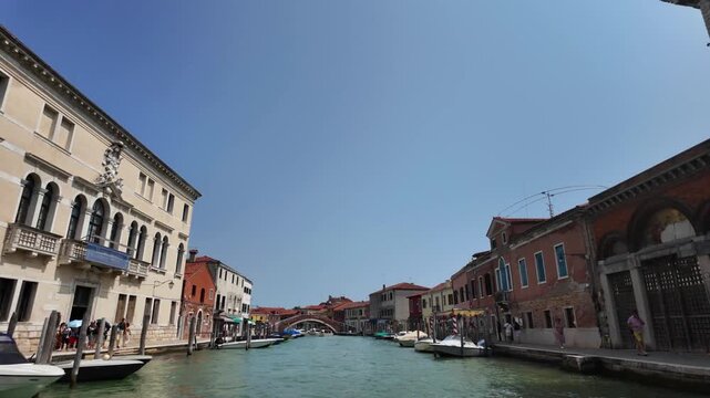 View of Venice from a boat slowly approaching across the lagoon. On either side, old houses built close to the water, boats, and pedestrian walkways.