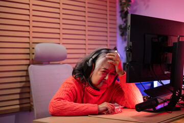 Obraz premium Sleeping senior eldery Asian woman with headphones streamer playing video game using desktop computer at gaming room at home. colorful neon background