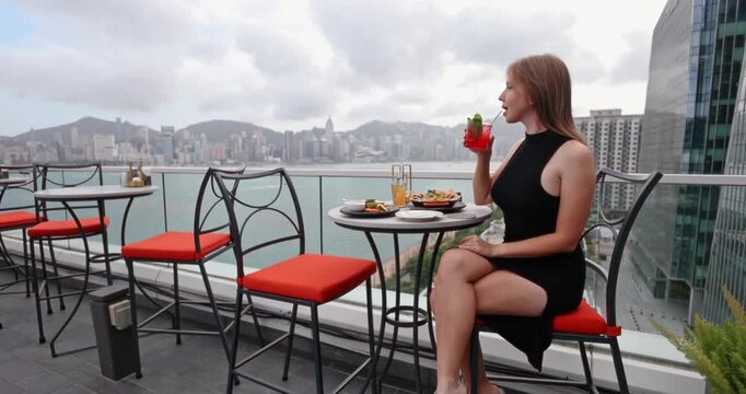Woman enjoying a refreshing cocktail at a luxurious rooftop bar, overlooking the panoramic Hong Kong city skyline and Victoria Harbour. Luxury travel, urban lifestyle, relaxation, fine dining