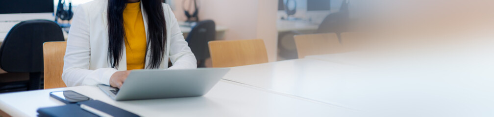 Asian businesswoman using laptop with large copy space on right side. Professional female worker in white blazer working at desk, office banner for marketing and website header. © Thawatchai Images