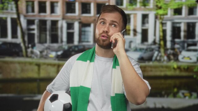 Man holding soccer ball with phone to ear on street beside building in amsterdam; match day excitement.