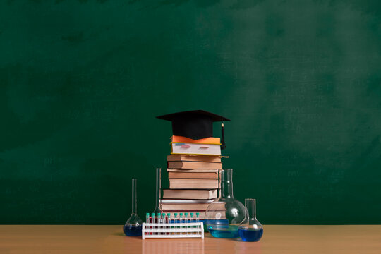 Education concept from school to college and university, textbooks, chemistry glassware and mortarboard on table against chalkboard. Blank space, copy space.