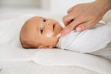 Infant with light brown hair lying on a soft surface while a hand applies cream to the baby's cheek in a bright indoor setting