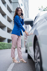 Fototapeta premium Confident Asian businesswoman smiling while on her smartphone, opening her parked car door as she prepares for a daily commute or business trip in the urban morning bustle