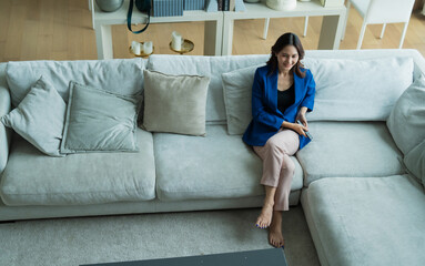 Obraz premium Woman sitting barefoot on a comfortable sofa in a modern living room, holding a remote control and enjoying an evening of television viewing, finding leisure and comfort alone