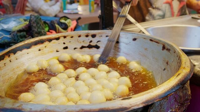 Street vendor frying round dough balls zalabia in hot oil inside metal pan. Vendor uses metal tongs to turn golden pieces of lugaimat while oil bubbles around them.Khan El Khalili bazaar, Cairo, Egypt