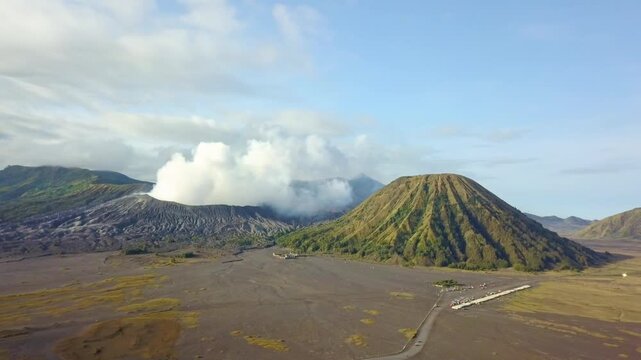 Stunning aerial view of Mount Bromo emitting white smoke alongside Mount Batok in East Java, Indonesia. Majestic volcanic landscape at Bromo Tengger Semeru National Park during a clear morning.