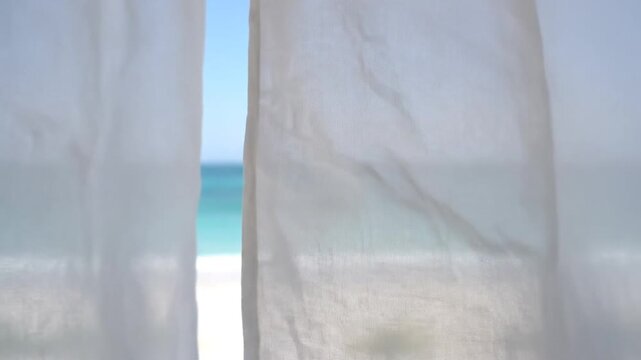 View of a beach scene through white curtains, revealing blue sky, ocean waves, sandy shore, and distant horizon with calm water and light breeze.