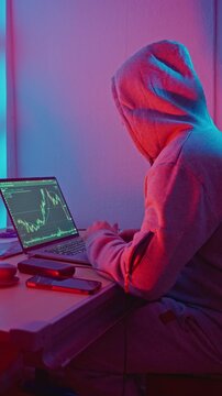 Caucasian Man In Hoodie Studying Charts At Laptop, Focused Analyst Reviewing Trend Lines And Indicators, Side Profile At Desktop Setup With Phone And Tools, Latenight Research Mood