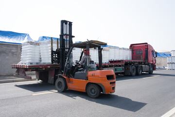 Orange forklift loading white bagged goods onto flatbed truck at industrial site
