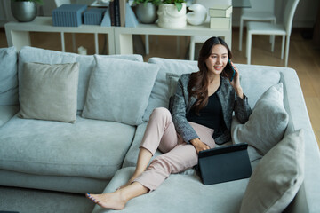 Obraz premium Woman in smart casual attire comfortably working from home, making a business phone call while sitting on a sofa with a tablet beside her, representing flexible modern work