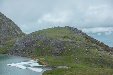 Naklejka premium Gloomy view to turquoise lake near green hill in hanging valley against misty hills in poor visibility in low clouds. Ice floes float in upland lake in mountain cirque against foggy rainy vastness.