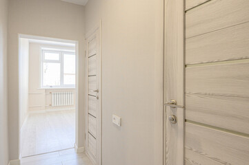 A bright hallway with light wood-finish doors, beige walls, light flooring, leading to a room with a window and white radiator