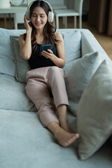Fototapeta premium Young Asian woman smiling, listening to music with headphones, and browsing her smartphone while relaxing comfortably on a living room sofa, enjoying leisure time at home
