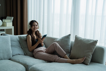 Obraz premium Young Asian woman smiling on a cozy sofa, wearing wireless headphones and holding a smartphone while streaming music and relaxing in her modern living room at home