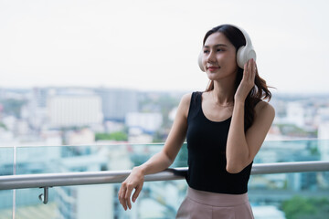 Stylish young woman wearing headphones listening to music, standing on a modern balcony with a...