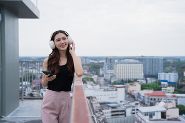 Fototapeta premium Young woman standing on a city rooftop, wearing headphones, holding a smartphone, and smiling while listening to music with urban buildings in the background