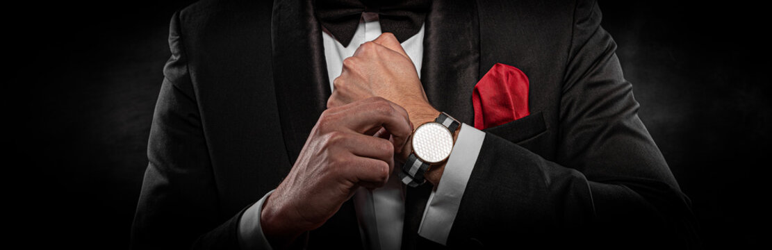 A man in a formal black tuxedo and bow tie adjusts a stylish wristwatch with a striped strap against a dark background.