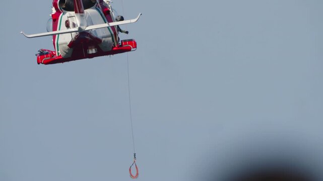 LUQA, MALTA - SEPTEMBER 28, 2025: A red and white search and rescue helicopter hovers in the sky. A crew member is visible in the open door as a rescue hoist line with a sling dangles below, preparing