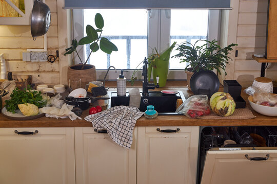 Messy modern kitchen after cooking with dirty dishes, fresh vegetables and cluttered countertop. Open dishwasher and black sink in realistic home interior. Everyday domestic life and household chaos 
