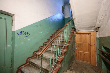 A worn Soviet-style staircase with green-painted walls, metal railing, brick steps, wooden door, graffiti, peeling paint, and a dimly lit communal hallway © evgeniykleymenov