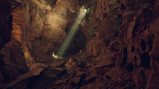 A stunning cavern features a beam of sunlight breaking through the rocky ceiling, illuminating the dark, rugged landscape below with a warm glow. The atmosphere evokes mystery and wonder.