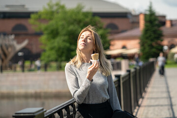 Fototapeta premium Carefree positive business woman enjoying sunny weather and ice cream, walking outdoors on summer day standing on bridge eating snack and squinting at bright sun, female freelancer on break in park.