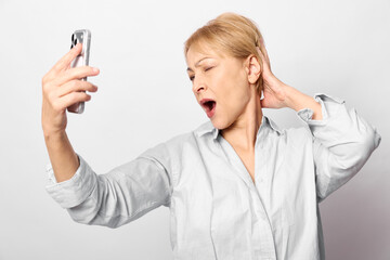 Fototapeta premium Yawning middle aged woman in casual shirt takes selfie on smartphone against white background. Concept of tiredness, fatigue, and everyday life moments captured.