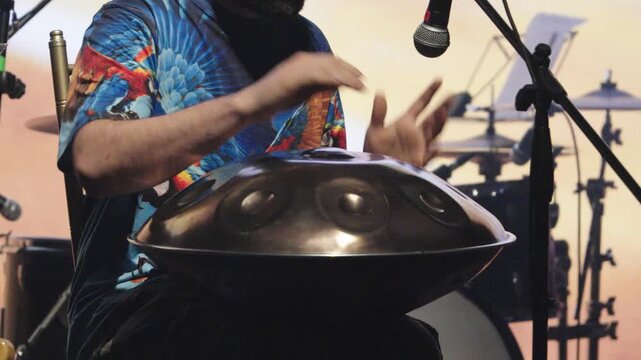 Horizontal medium shot of a musician performing with a handpan (hang drum) on stage. The artist wears a vibrant t-shirt with a blue macaw print.