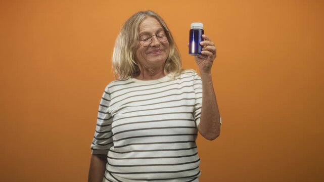 Woman holds blue pill bottle in a raised hand while smiling and examining the label against an orange studio wall; wellness hope confidence.