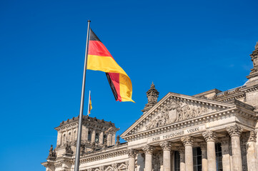 Obraz premium Berlin, germany, august 15, 2023. The german national flag waving in front of the historic reichstag building under a clear blue sky, symbolizing government and democracy