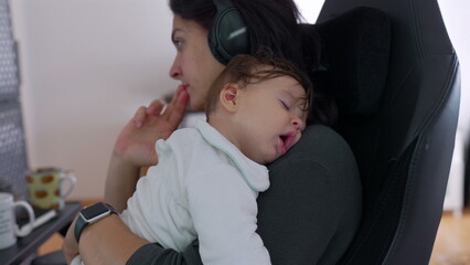 Mother working while holding sleeping baby with mouth slightly open, resting peacefully on her...