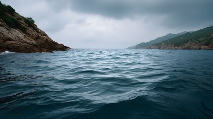 A dramatic view of a choppy dark blue sea between rocky cliffs and green hills under a cloudy sky