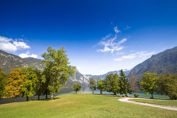 Beach area of Stara Fuzina on Lake Bohinj (Bohinjsko Jezero), Slovenia, on a sunny afternoon, with green lawn, calm water, and the Julian Alps mountain landscape under a clear blue sky.