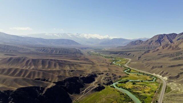 Aerial view captures winding Chu river flowing through vast valley surrounded by barren mountains and snow-capped peaks. Serene landscape showcases natural beauty and remote tranquility.