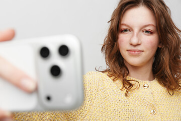 Young woman with wavy brown hair wearing yellow sweater takes selfie with smartphone on white...