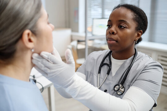 Young adult Black female doctor examining face of Caucasian middle aged woman patient in medical office, wearing gloves and stethoscope, performing professional healthcare assessment