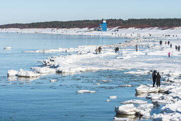 Icy winter seascape with frozen waves, floating ice floes on the blue sea water along a snowy shoreline and people walking on the Baltic Sea in Palanga, Lithuania