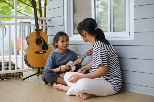 Two young Asian sisters and siblings playing ukulele together at home. Older sister teaching music on a musical instrument. Concept of happy family relationship, education, and lifestyle.