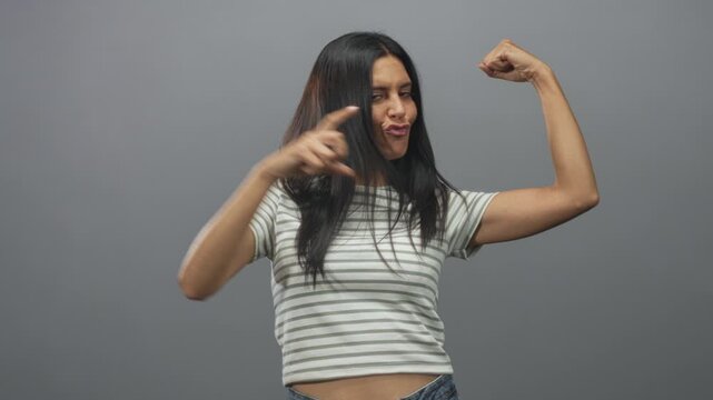 Young woman flexes bare arms showing biceps and points finger to muscle while making playful pout in studio grey backdrop; confidence empowerment.